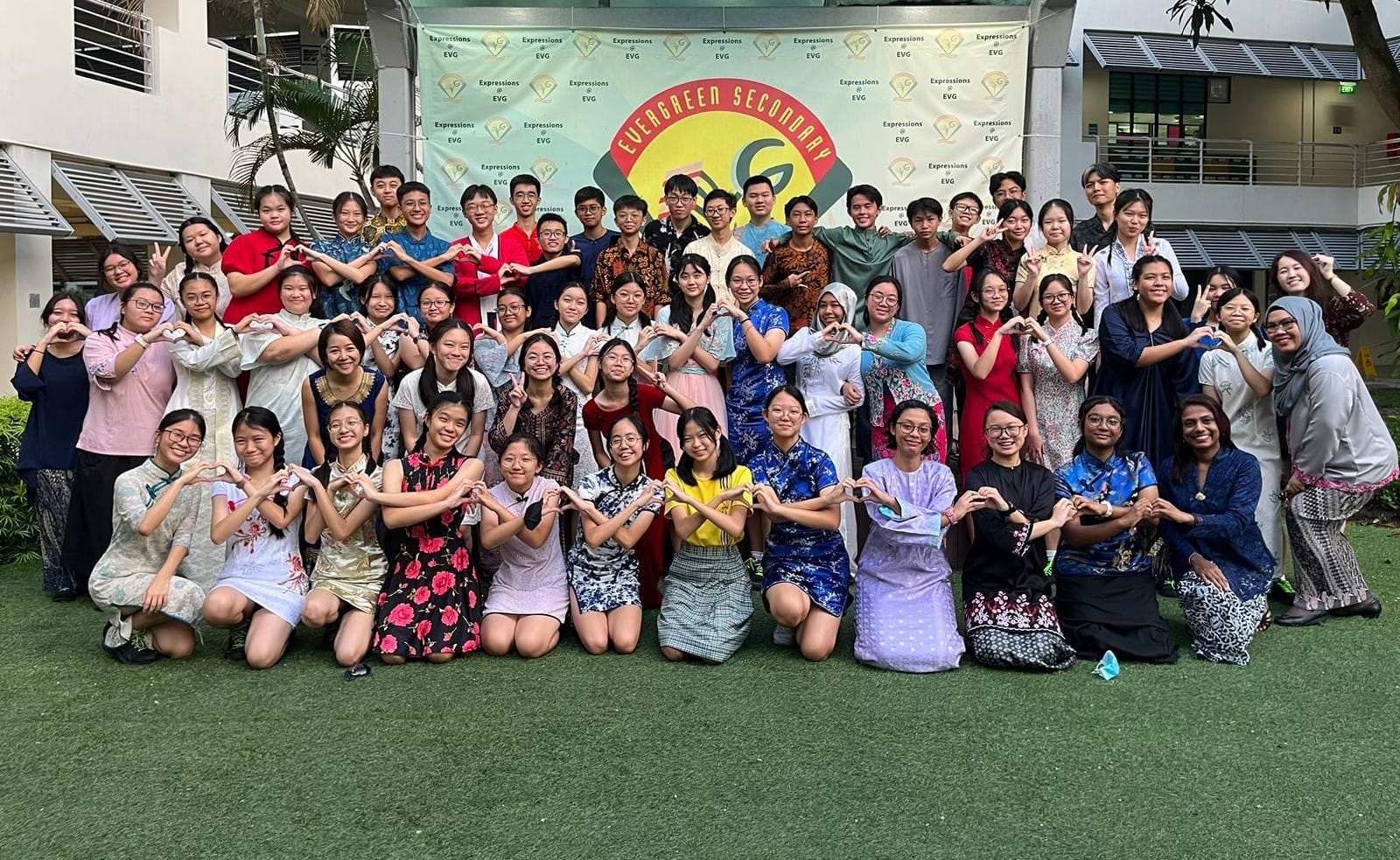 Large group of students in cultural attire making heart shapes with hands in front of Evergreen Secondary School banner.